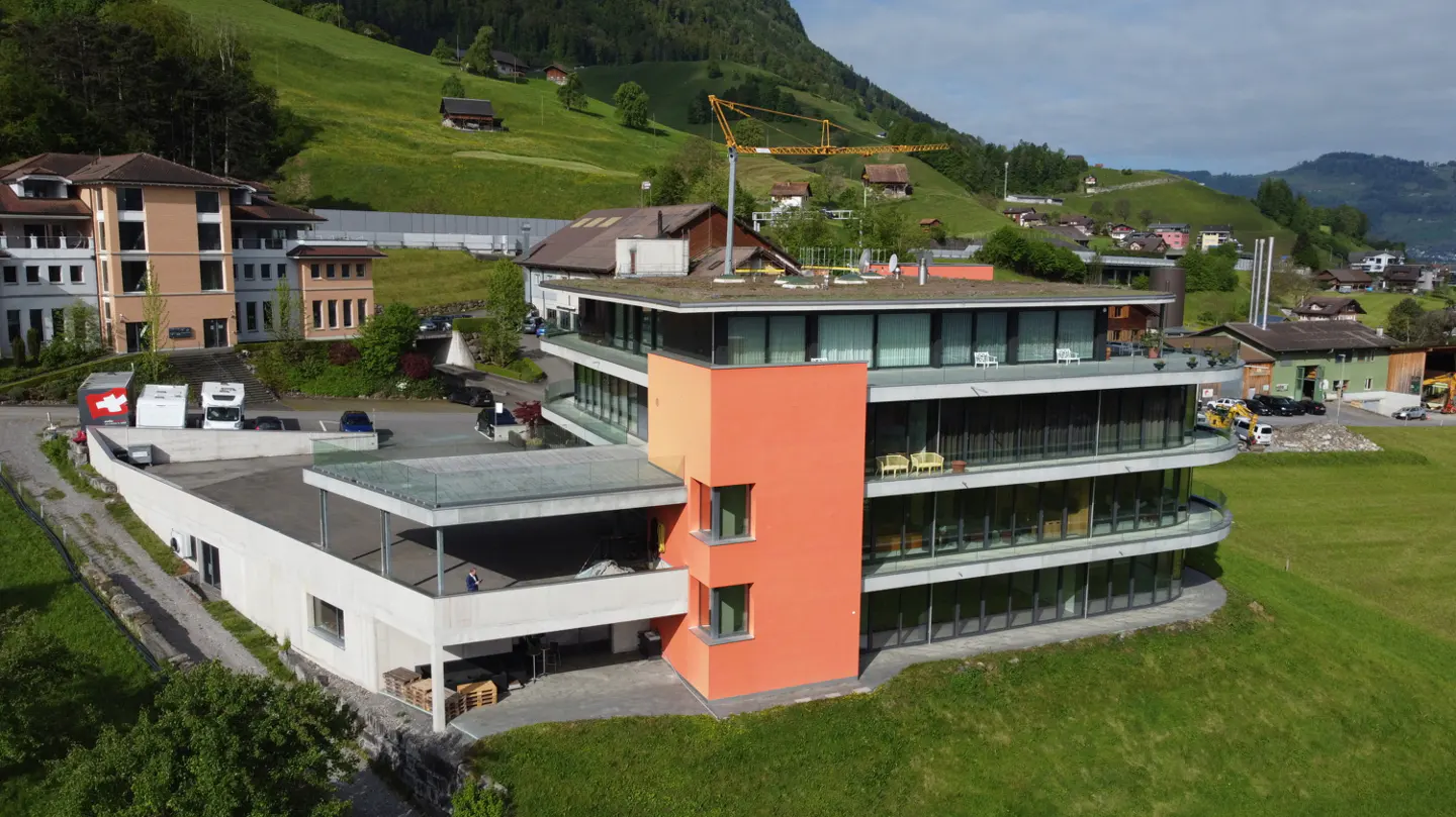 Modern building with orange accent, surrounded by green hills and other buildings in a rural setting.