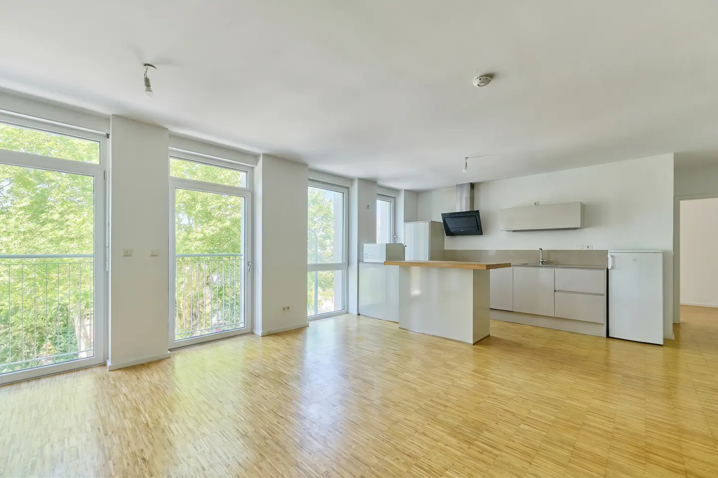 Bright open-plan kitchen and living area with light wood floors, white cabinets, and floor-to-ceiling windows with trees.