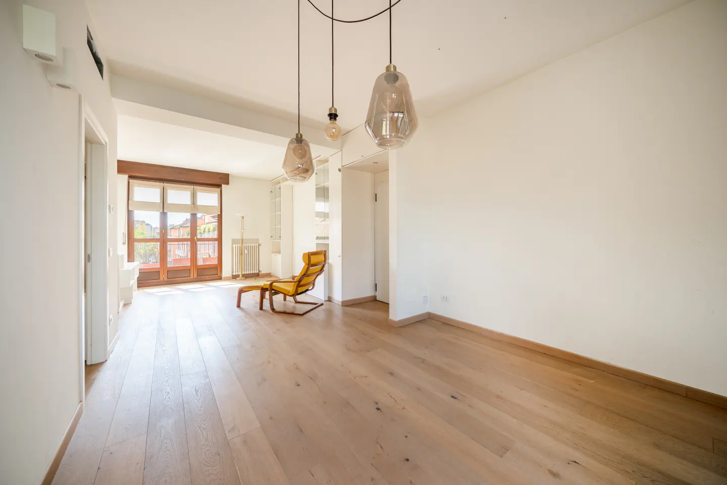 Bright, empty room with wood floors, white walls, and three pendant lights. A yellow lounge chair sits near a window with a view.