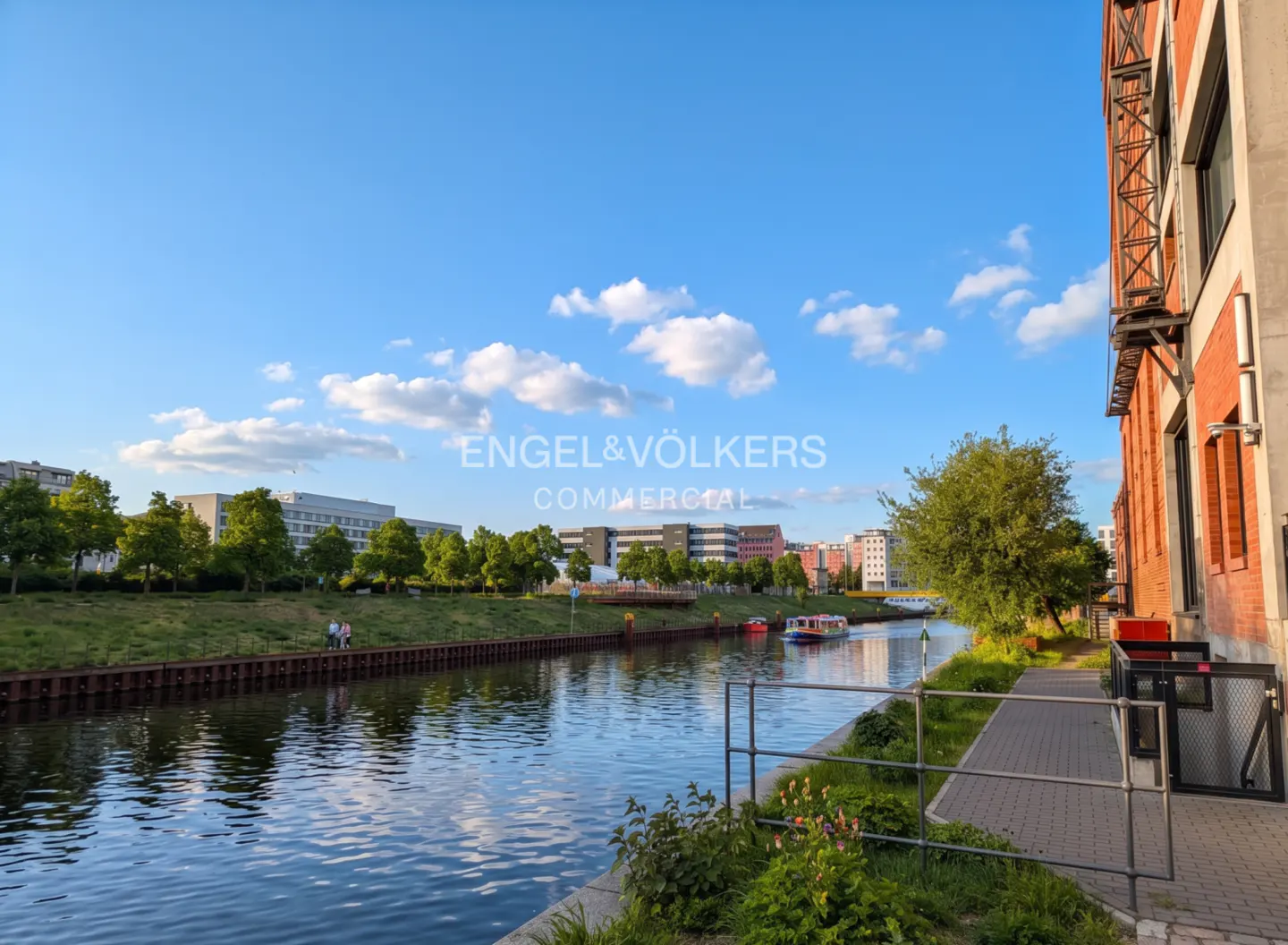 Scenic view of a canal with a boat, green trees, and a brick building under a blue sky with clouds.