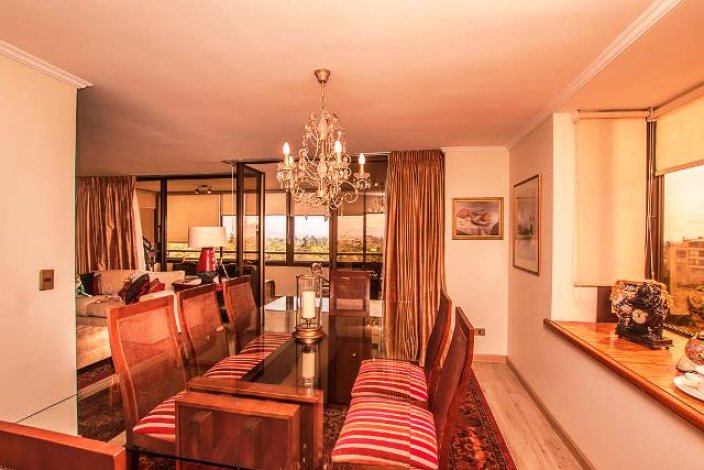 A dining room with a glass table, wooden chairs with striped cushions, and a chandelier. Curtains and a city view are visible.
