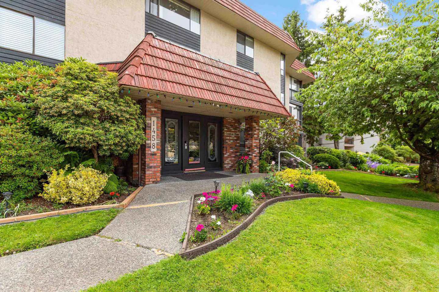 Exterior view of a multi-story apartment building with a red tile roof over the entrance. Lush green lawn and flower beds surround the walkway.