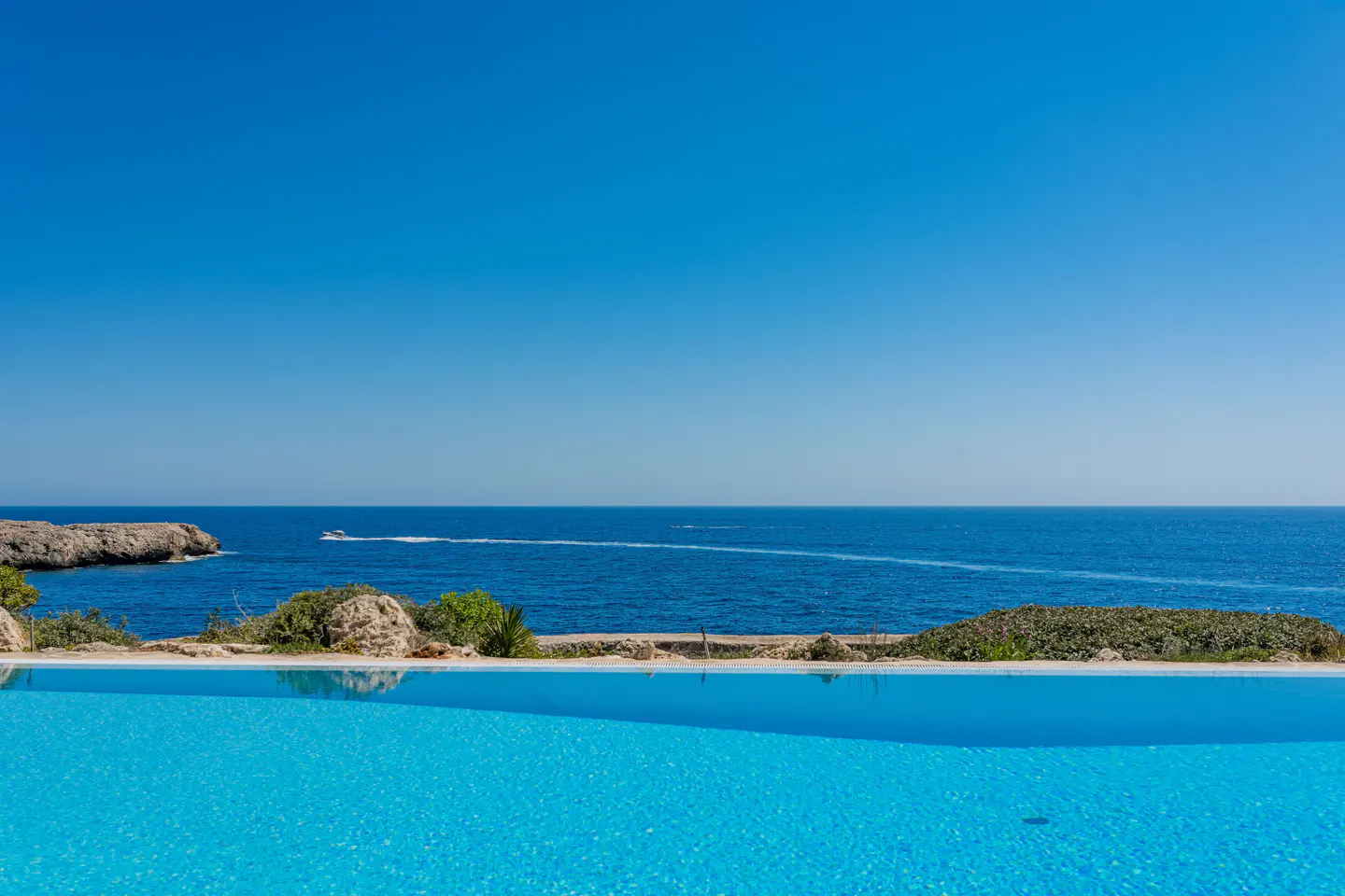 Infinity pool with turquoise water overlooking the blue ocean and sky. A boat is seen in the distance.