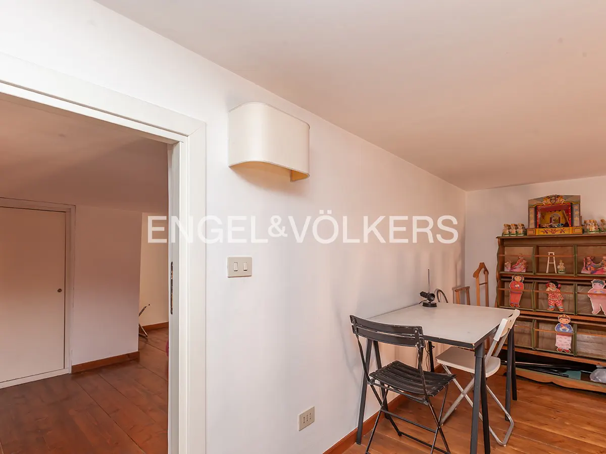 A bright room with white walls, a table with two chairs, and a wooden cabinet with figurines. The Engel & Völkers logo is on the wall.