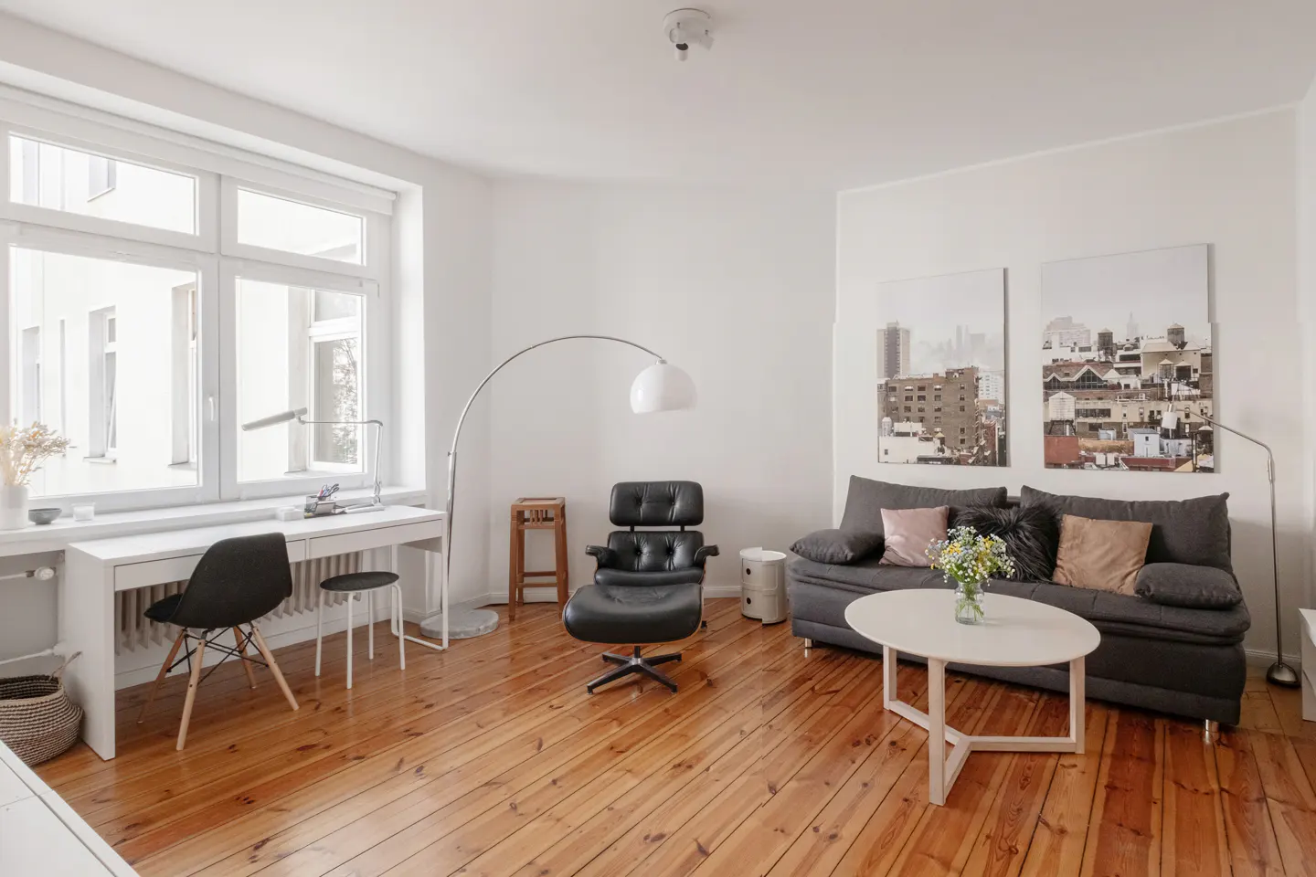Bright living room with wood floors, white walls, and large window. Features a desk, black chair, gray sofa, and black leather chair. Cityscape art on the wall.
