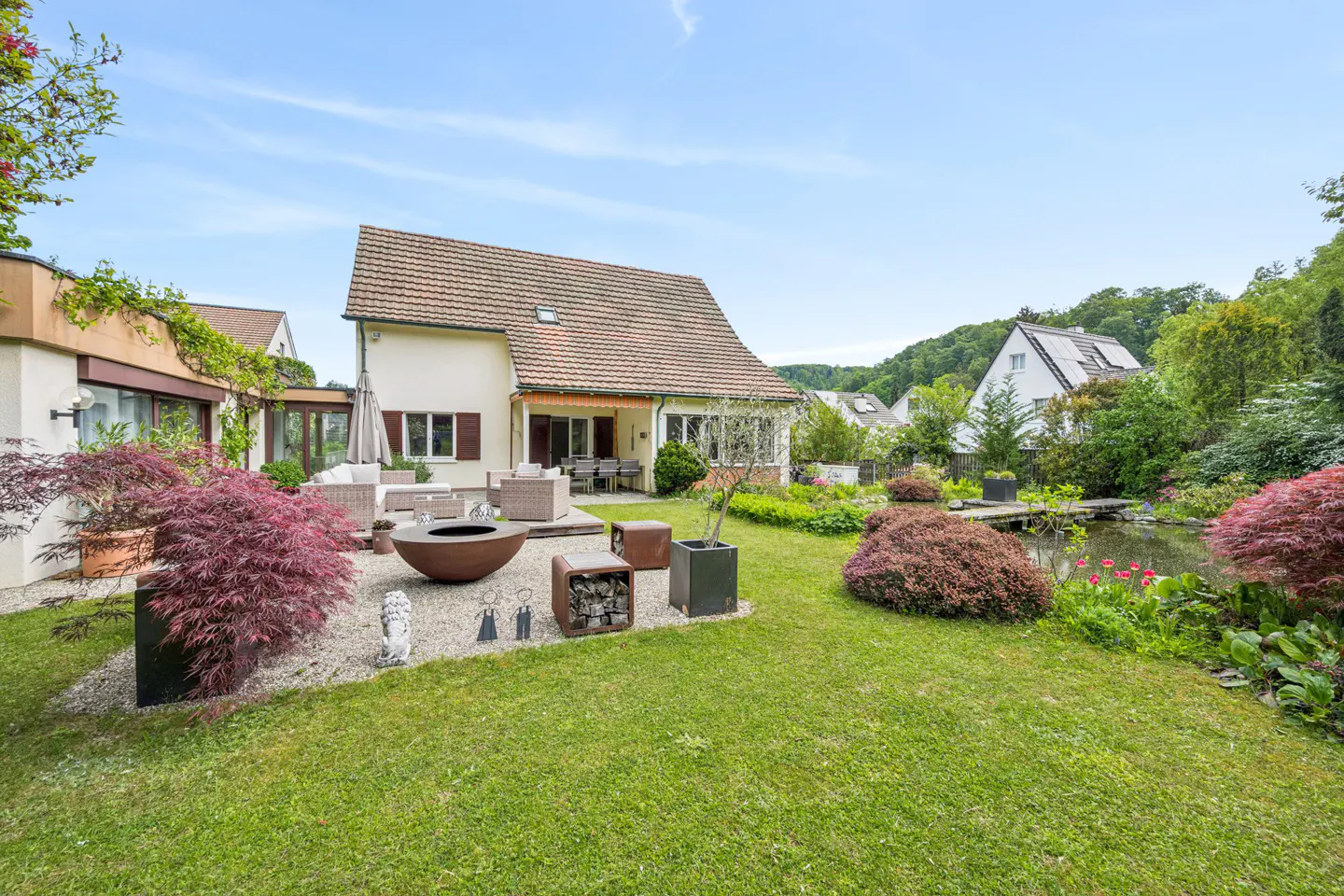 A house with a large green lawn, patio furniture, a fire pit, and a pond.