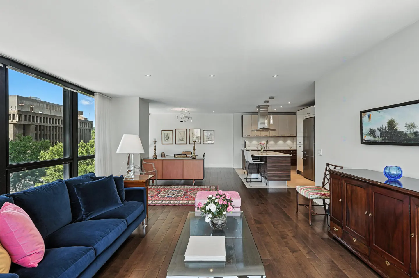 Bright, open-concept living room with hardwood floors. A blue sofa faces a large window with a city view. A kitchen is visible in the background.