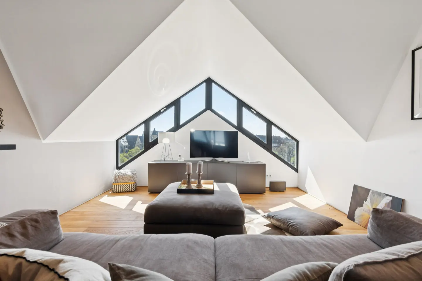 Modern attic living room with a large triangular window, grey sofa, ottoman, and TV on a console table.