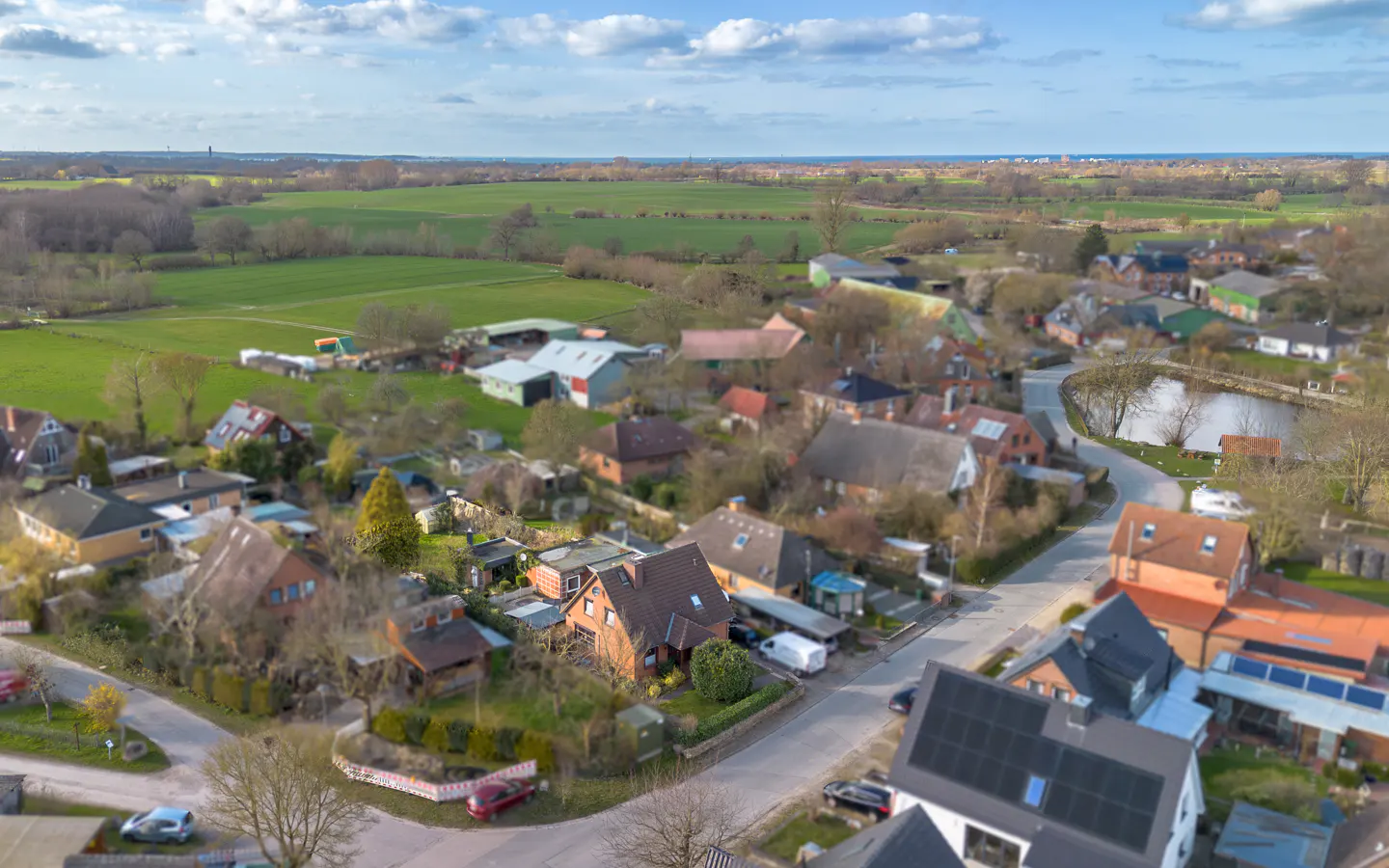 Aerial view of a village with houses, green fields, and a pond under a blue sky with clouds.