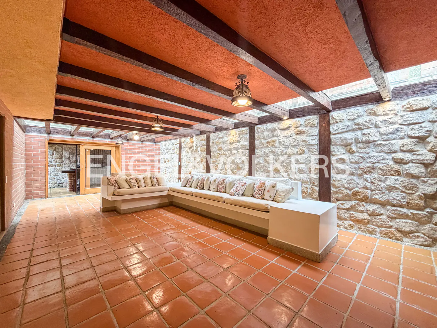 A sunroom with terracotta tile floors, stone walls, and a long white sofa with pillows. The ceiling has exposed wood beams and hanging lights.