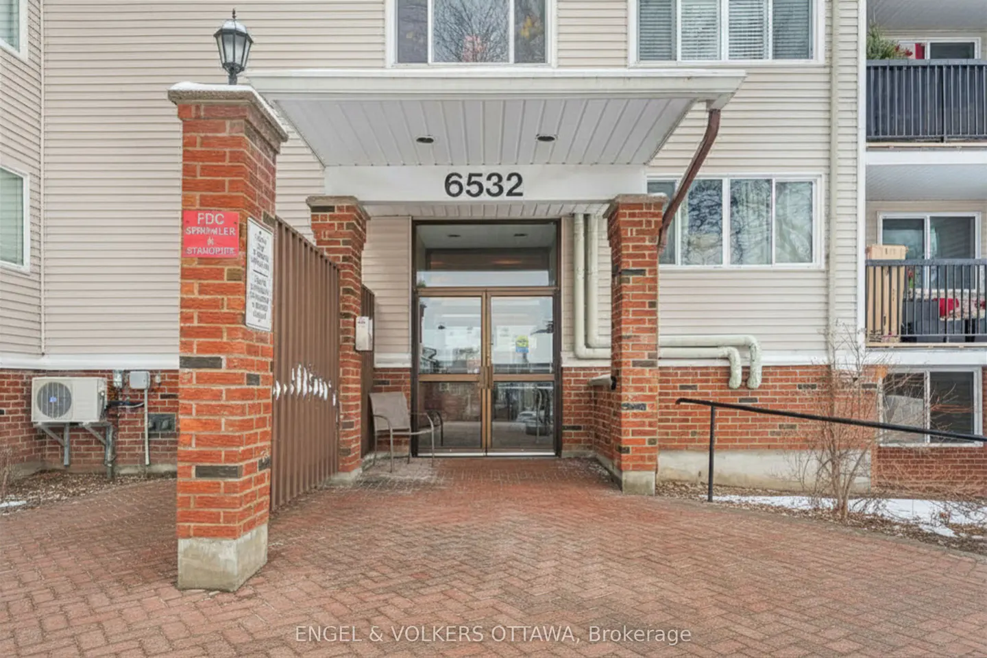 Exterior of a beige apartment building with the address 6532 above the glass double door entrance. A brick walkway leads to the entrance.