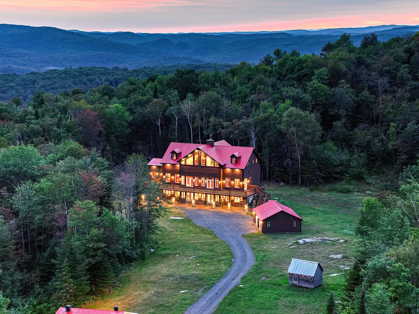 A large, brown house with a red roof sits atop a hill surrounded by green trees and mountains at dusk.