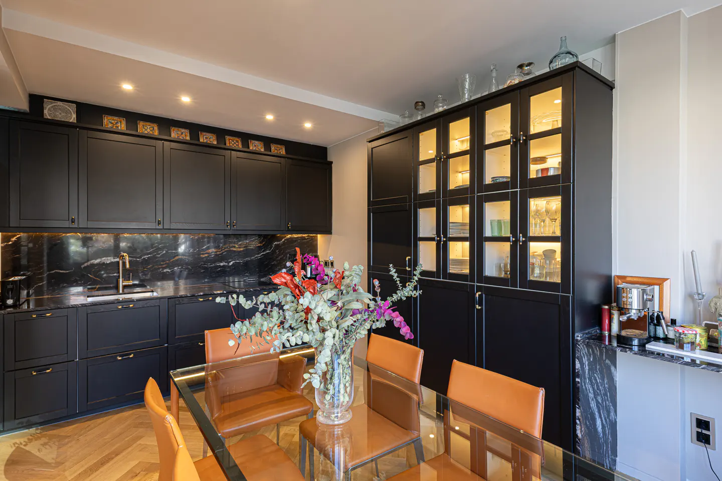 A modern kitchen with black cabinets, marble backsplash, and a glass dining table with orange leather chairs. A vase of flowers sits on the table.