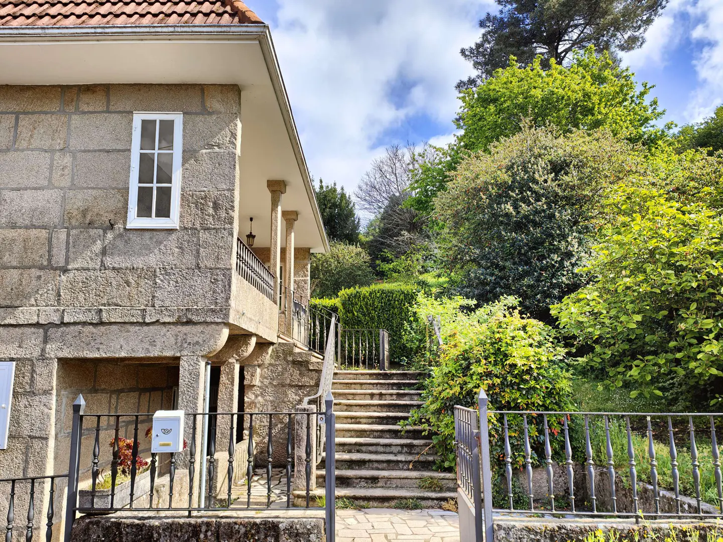 Exterior view of a stone house with a red tile roof, a balcony with columns, and a stone staircase leading up to the house, surrounded by lush greenery.