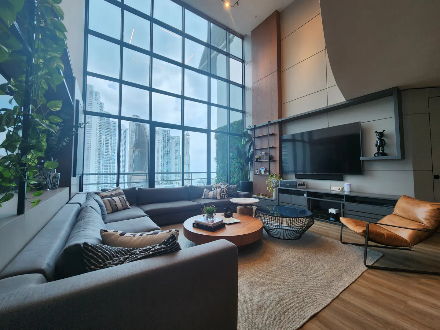 Living room with a gray sectional sofa, wood coffee tables, and a leather chair. Large windows offer a city view.