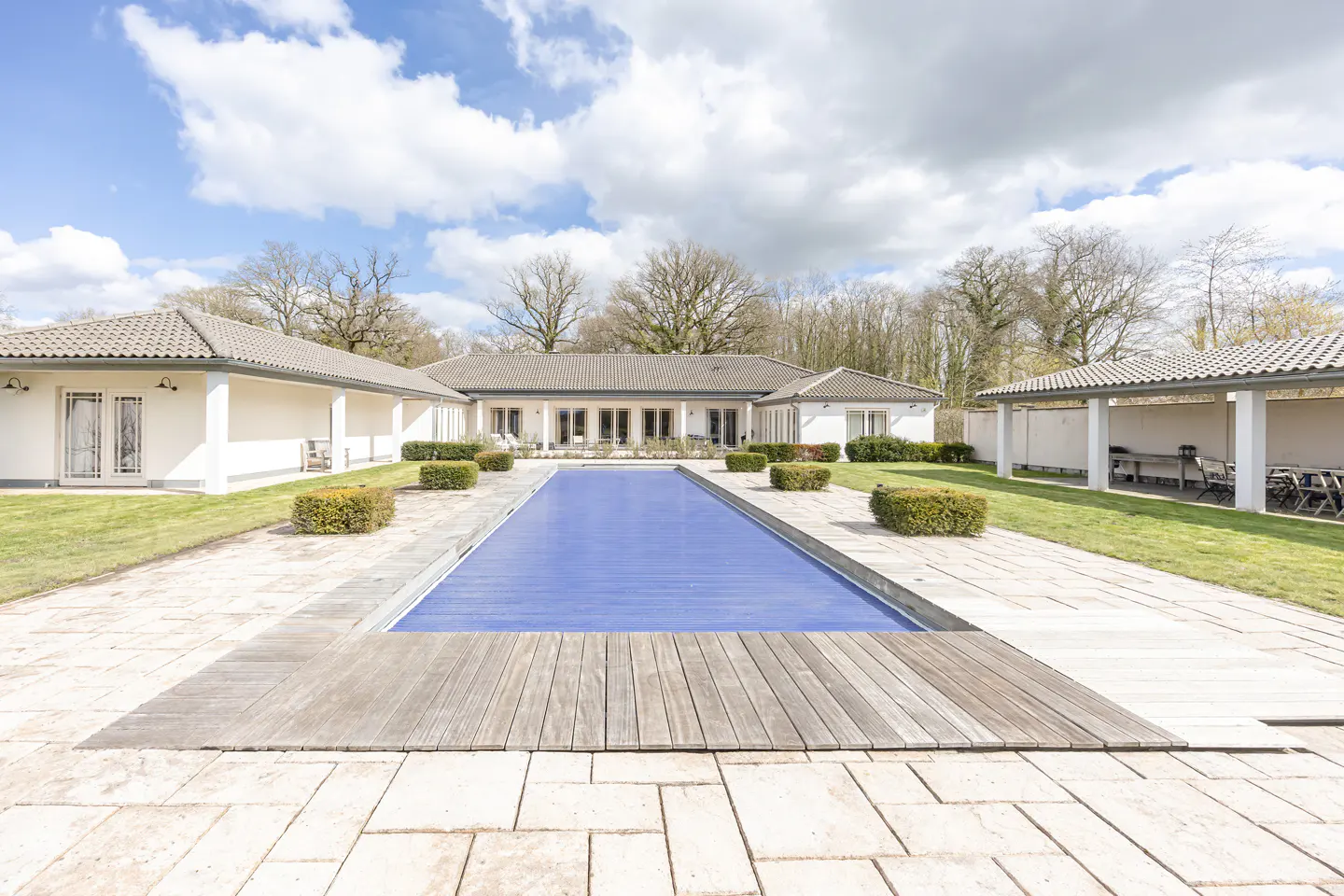 A wide shot of a covered blue swimming pool, surrounded by a white house, green grass, and trees.