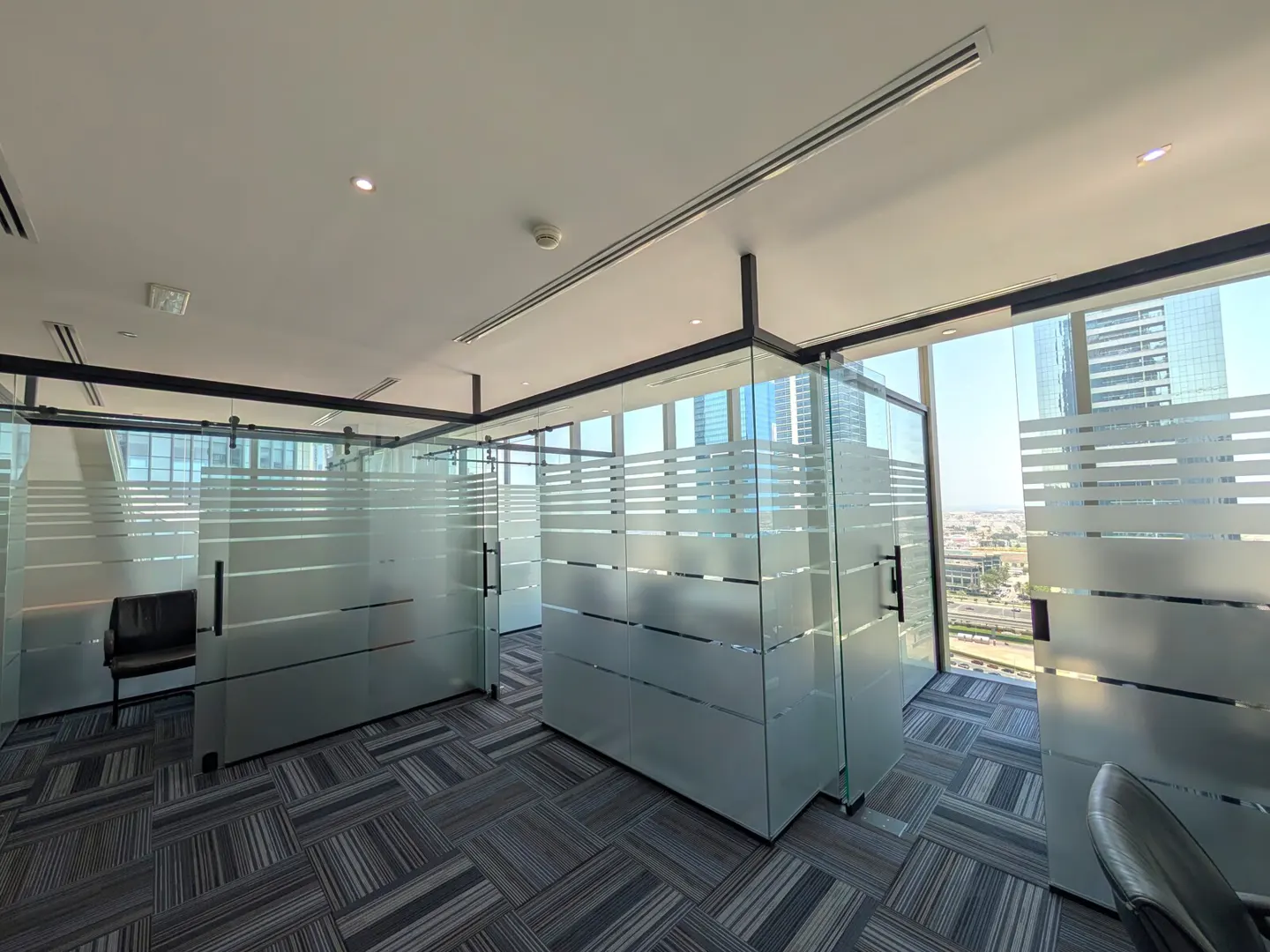 Office space with glass-walled cubicles, horizontal frosted stripes, and a patterned gray carpet. A city view is visible through the windows.