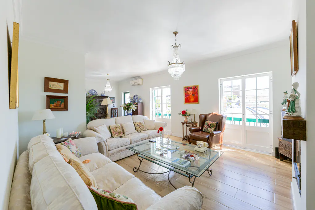 Bright living room with white walls, light wood floors, and a glass coffee table. Cream sofas, a brown leather chair, and crystal chandeliers add elegance.