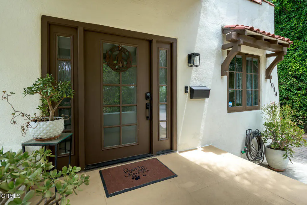 Front exterior of a white stucco house with a brown front door and window. A welcome mat and potted plants decorate the porch.