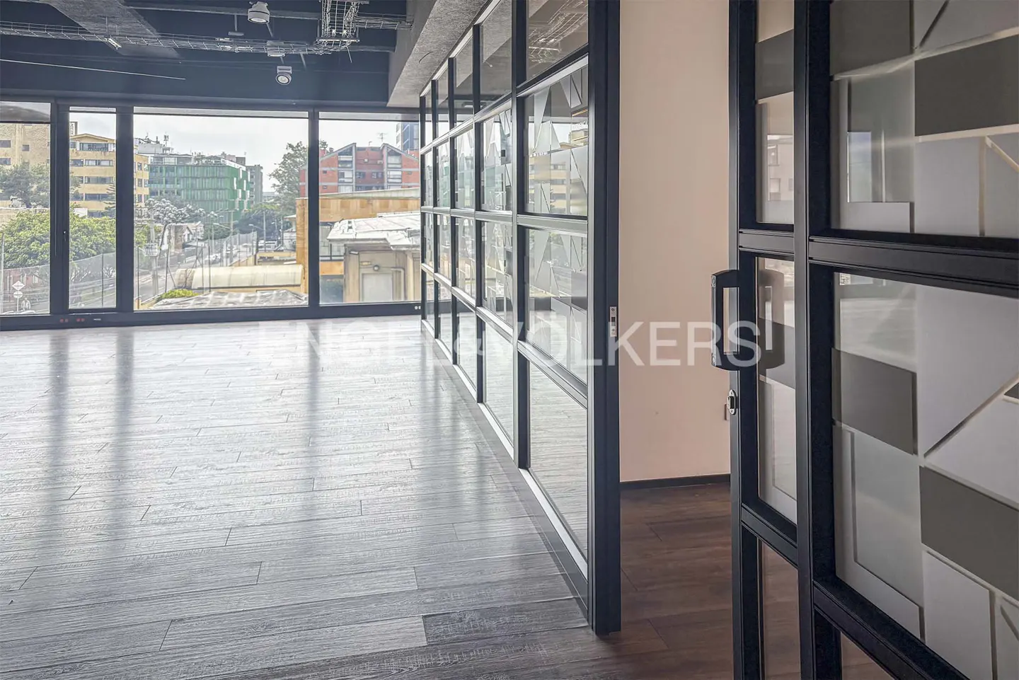 An empty office space with dark wood floors, large windows with a city view, and glass-paneled walls.