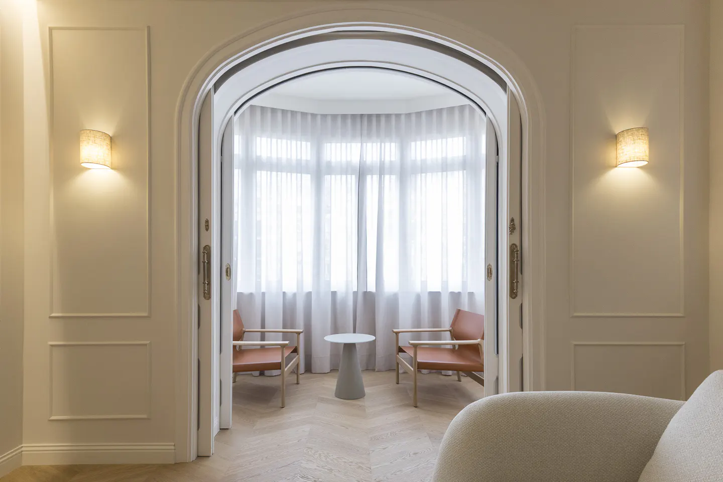 View through an arched doorway into a bright room with two orange chairs and a small table. White walls, sconces, and herringbone wood floor.
