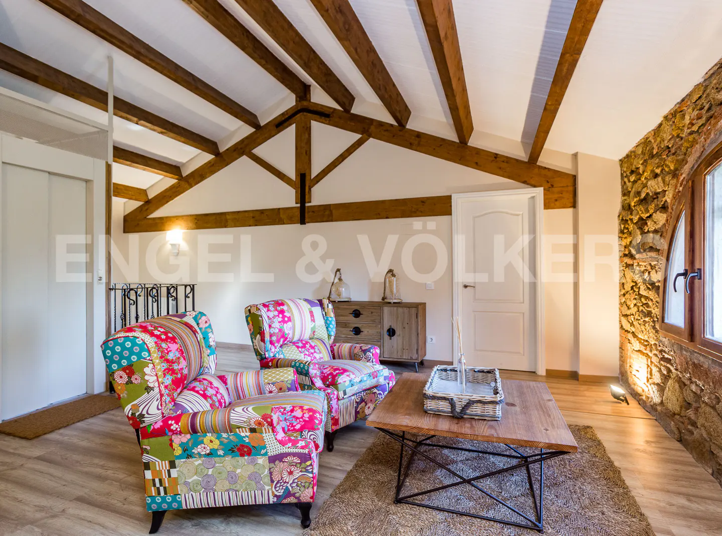 Living room with exposed beams, stone wall, and colorful patchwork armchairs. A wooden coffee table sits on a brown rug.