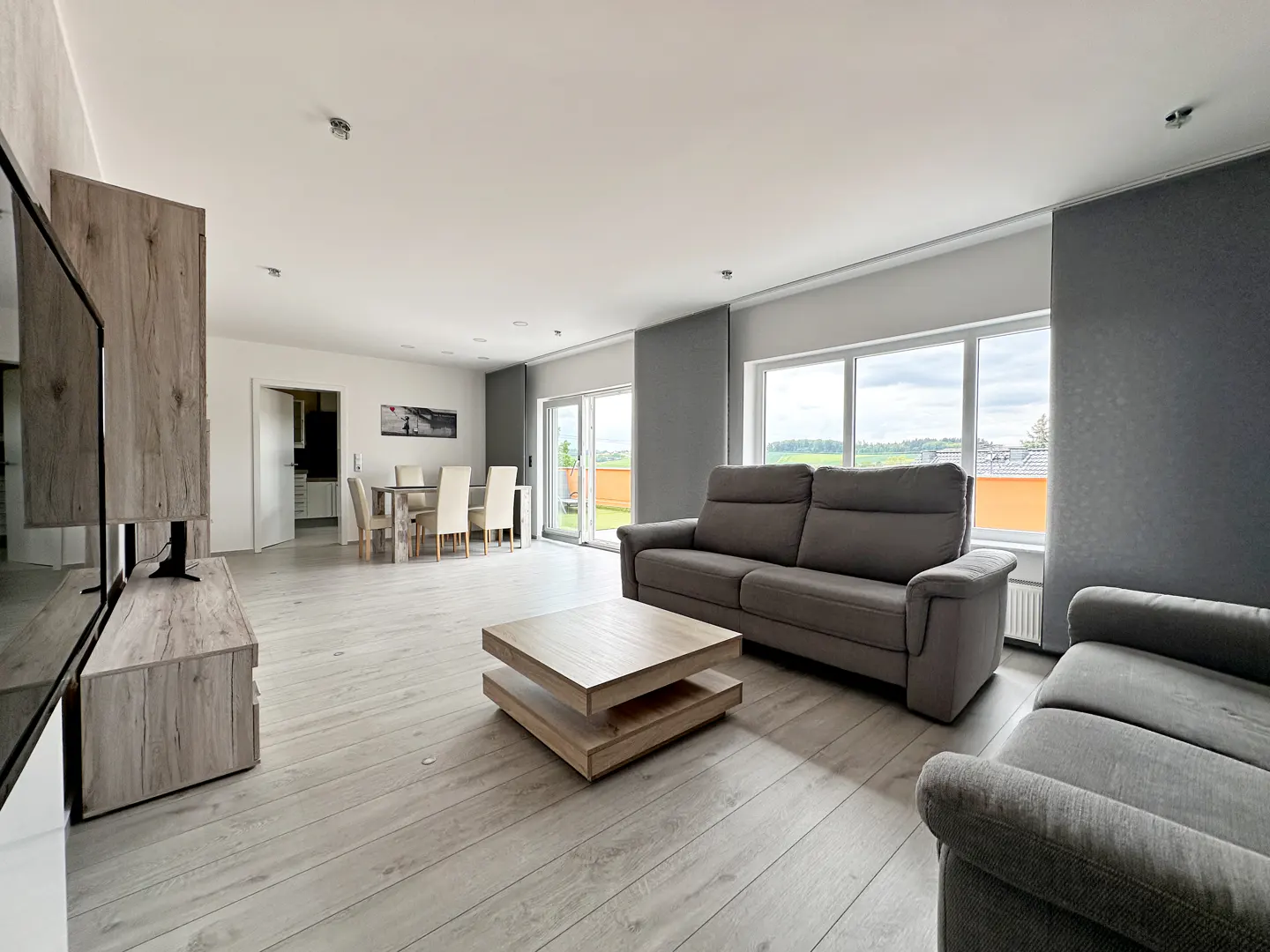 Living room with gray sofas, wood floors, and a light wood coffee table. Dining table and chairs are visible in the background.