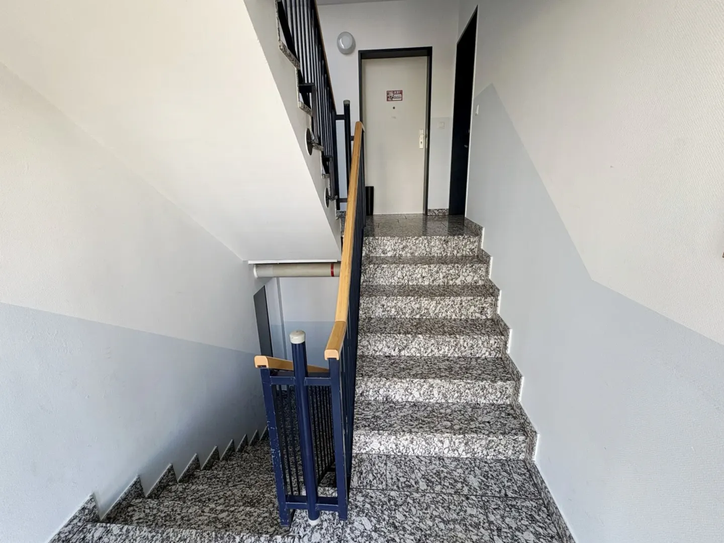 A stairwell with granite steps and blue railings leads to a white door on the upper level. The walls are painted white and gray.