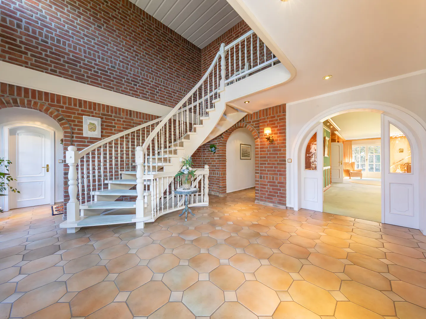 Bright foyer with a winding white staircase, red brick walls, and tan tile flooring. An arched doorway leads to another room.
