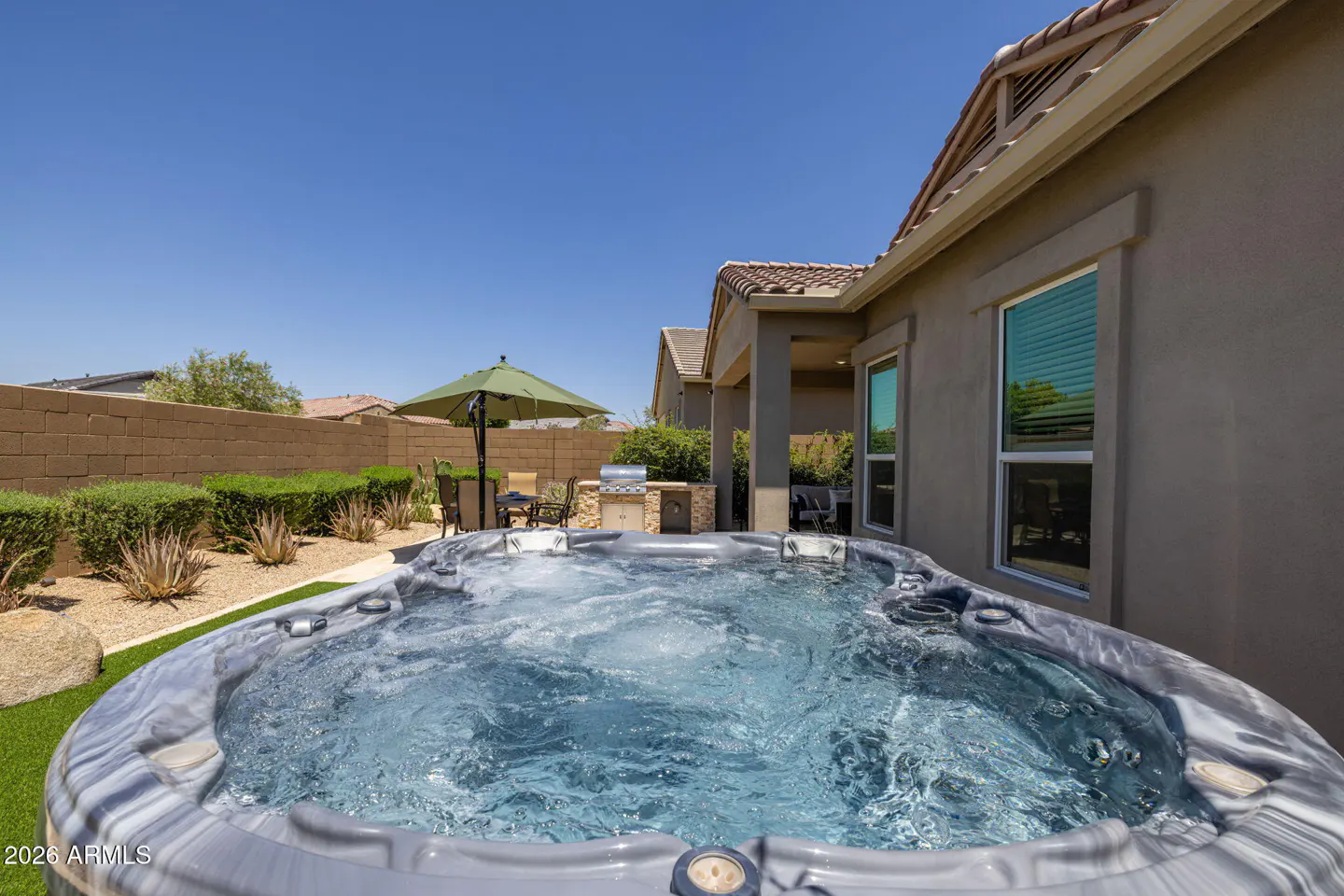 Outdoor spa with bubbling water, patio furniture, and a built-in grill against a tan house under a clear blue sky.