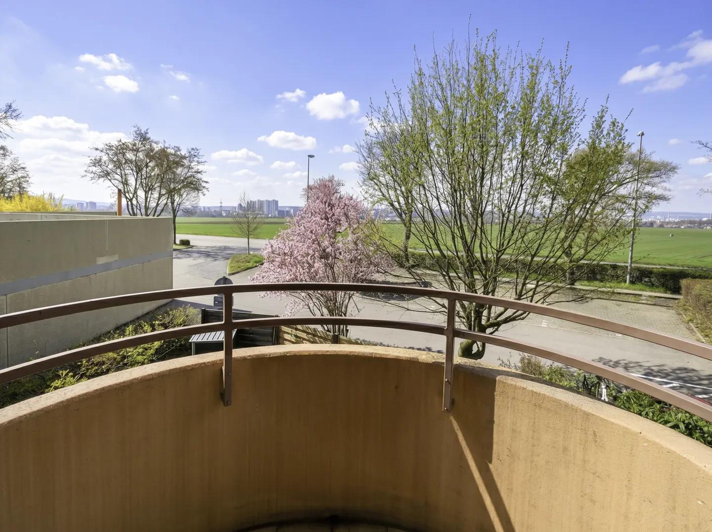 View from a balcony with a brown railing, overlooking a green field and trees under a blue sky.