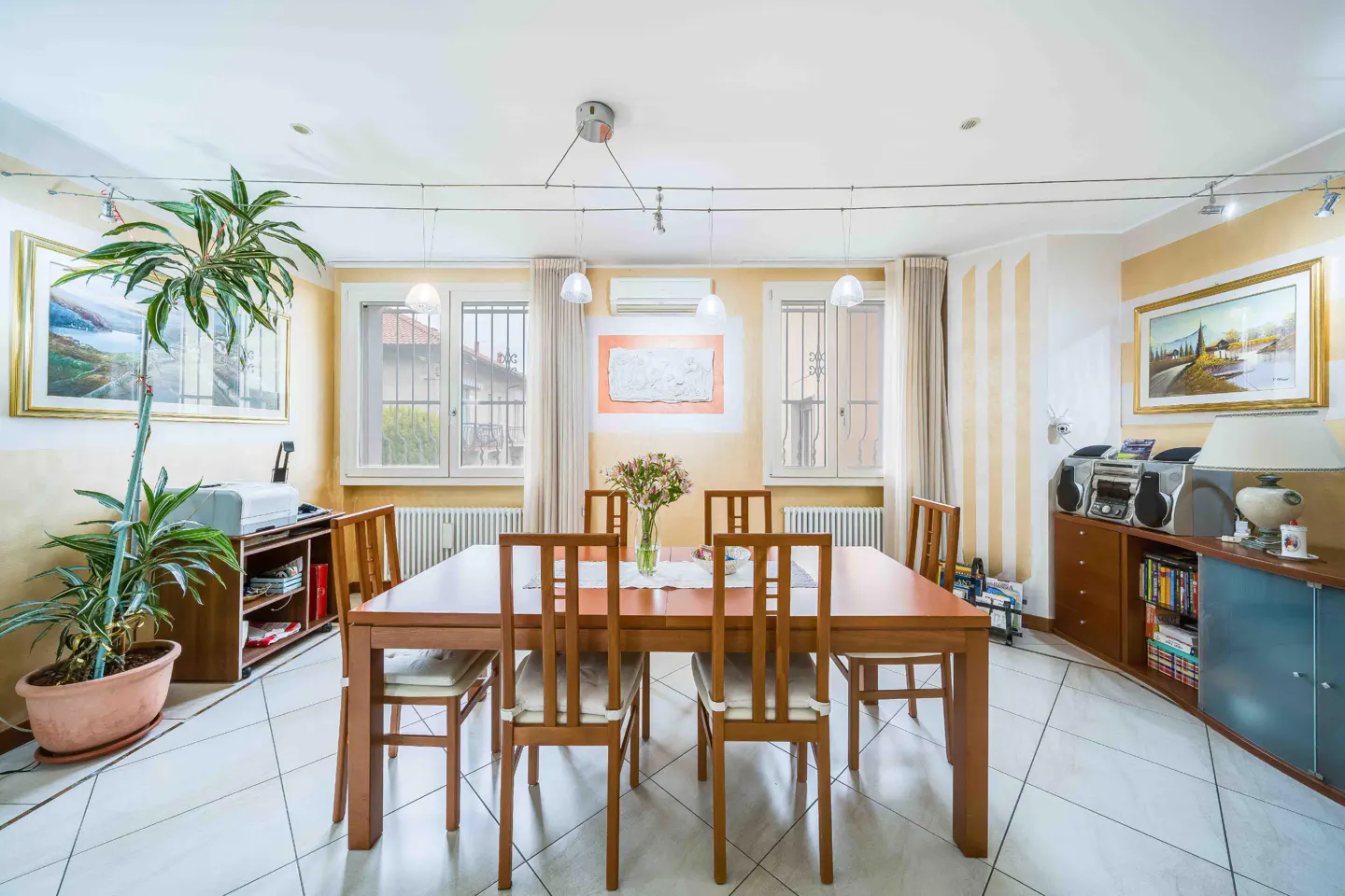 A bright dining room with a wooden table, chairs, and a vase of flowers. Windows with white curtains and artwork adorn the yellow walls.