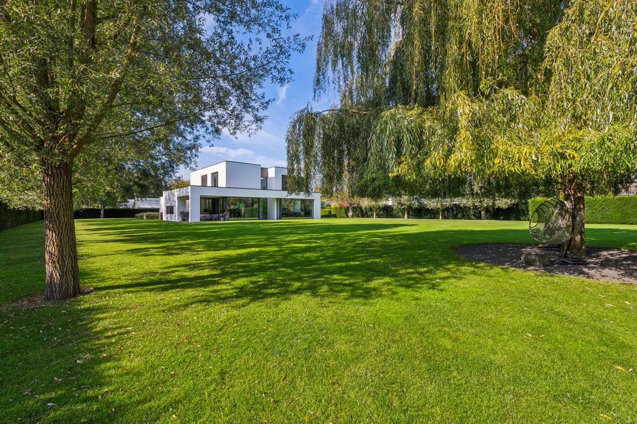 Modern white house with large glass windows, surrounded by a green lawn and trees under a blue sky.