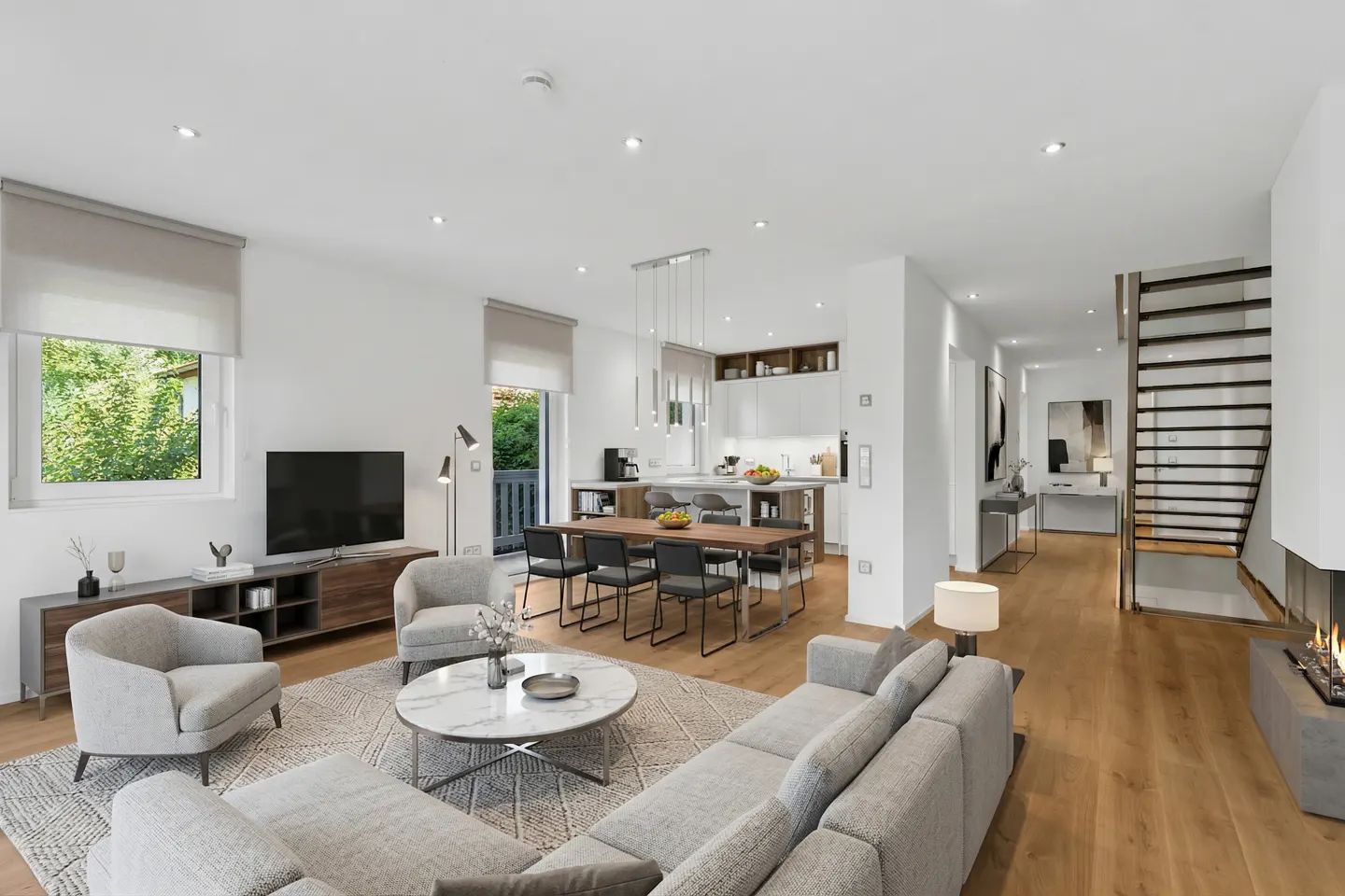 Bright, open-concept living space with gray sofa, chairs, and marble table. Kitchen, dining area, and staircase visible. Hardwood floors throughout.