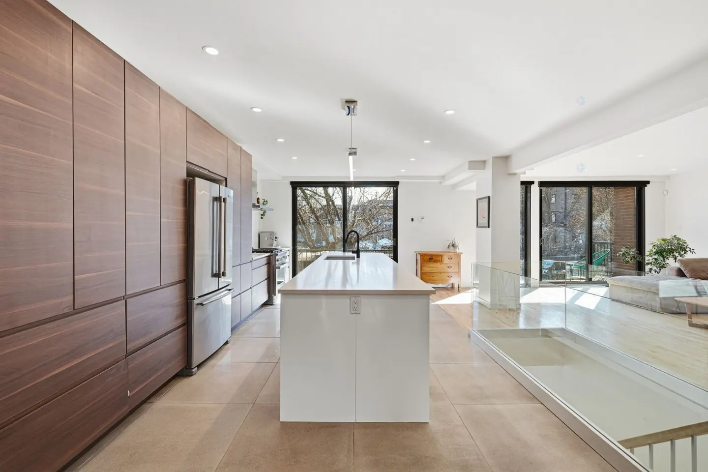 A modern kitchen with brown cabinets, a stainless steel refrigerator, and a white island with a black faucet. A glass railing separates the kitchen from the living room.