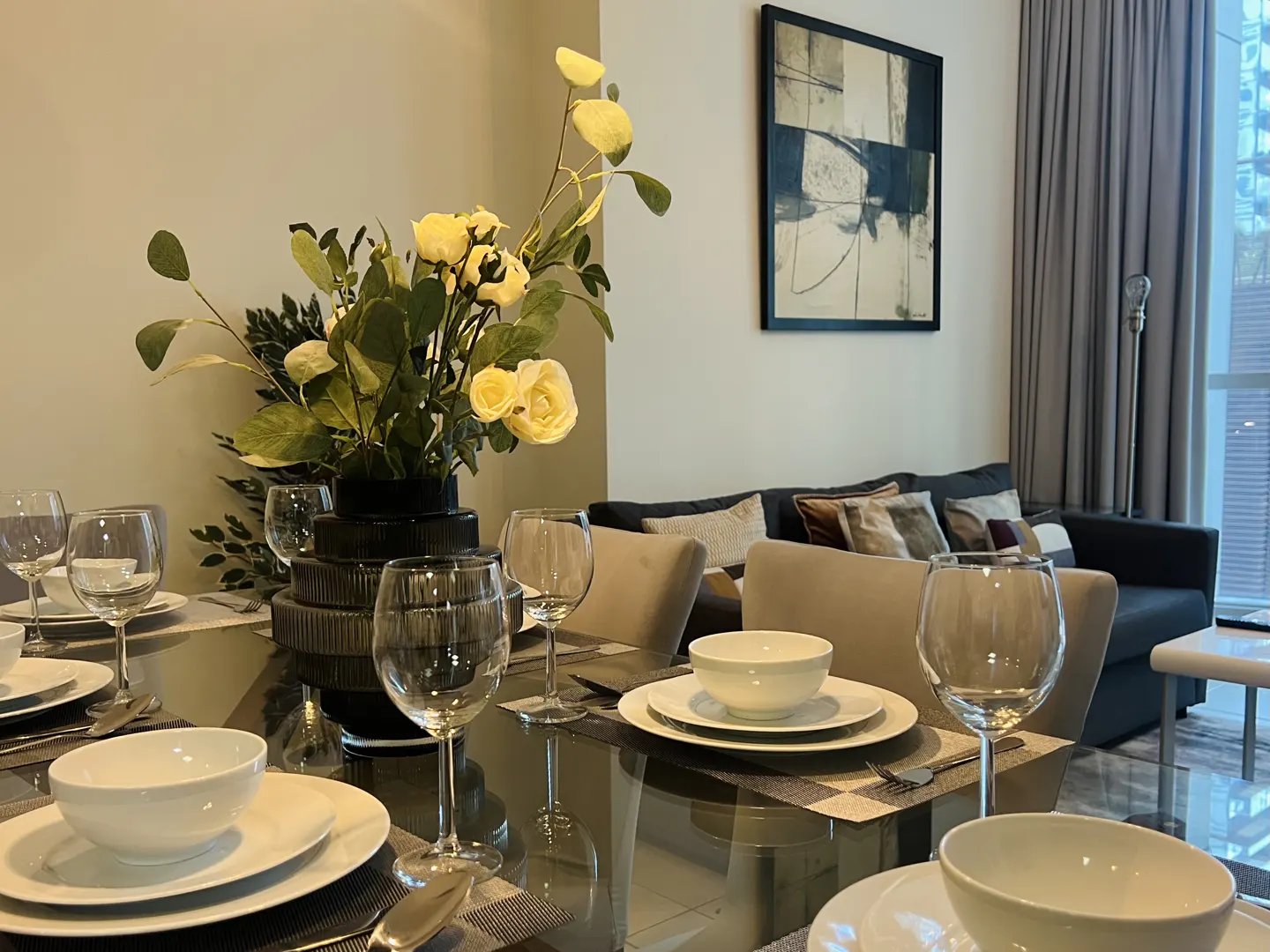 A dining table set with white plates, bowls, and wine glasses, with a black vase of yellow flowers in the center. A gray sofa is in the background.