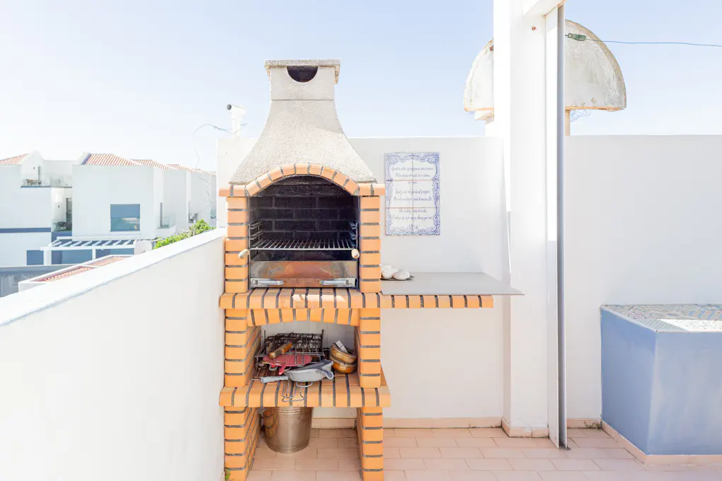 Outdoor brick barbecue with a chimney, metal grill, and utensils on a patio with white walls and a blue tiled bench.