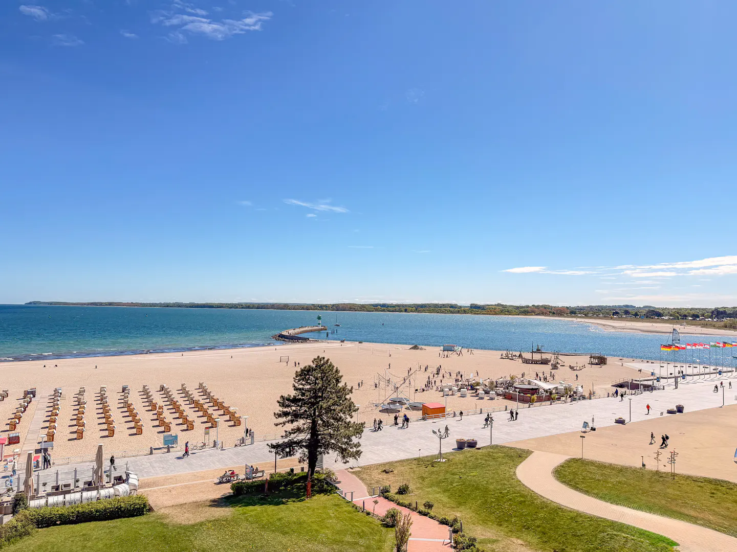 Beach scene with blue sky, turquoise water, and tan sand. Beach chairs line the shore, and people stroll along the boardwalk.