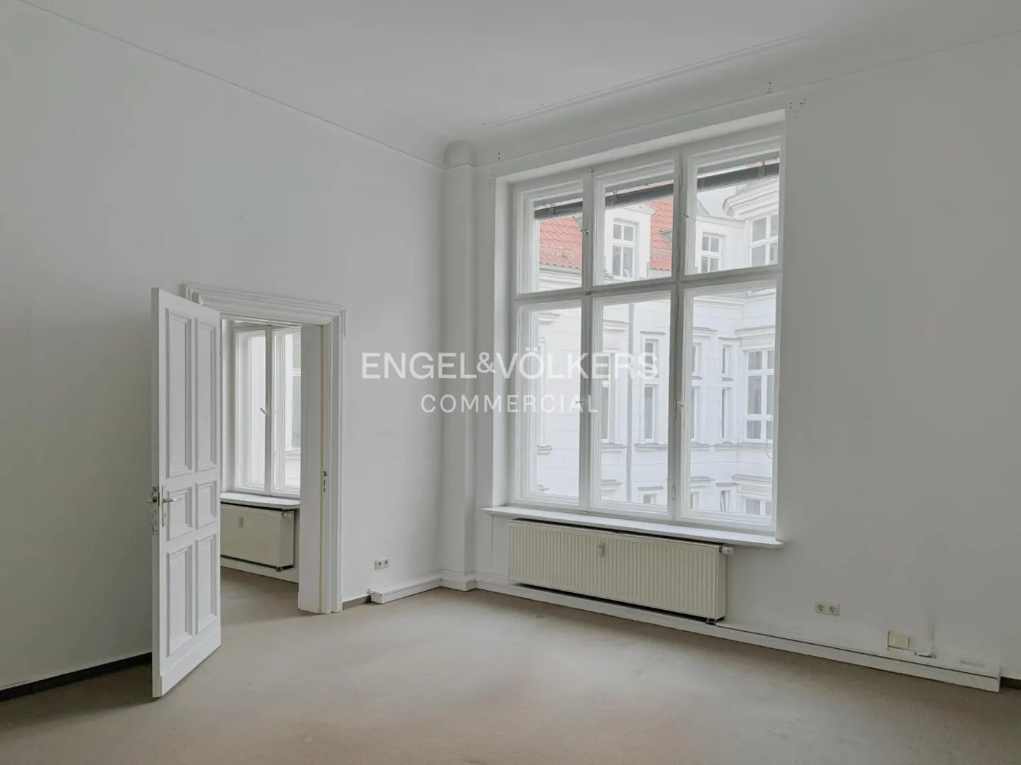 Bright, empty room with white walls, a large window, and an open white door. A radiator sits under the window. The Engel & Völkers logo is visible.