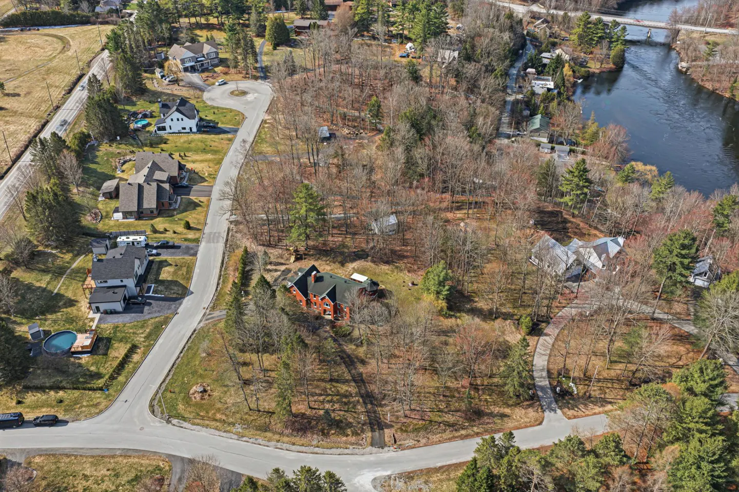 Aerial view of a residential area with houses, trees, and a river. A red brick house is nestled among bare trees.