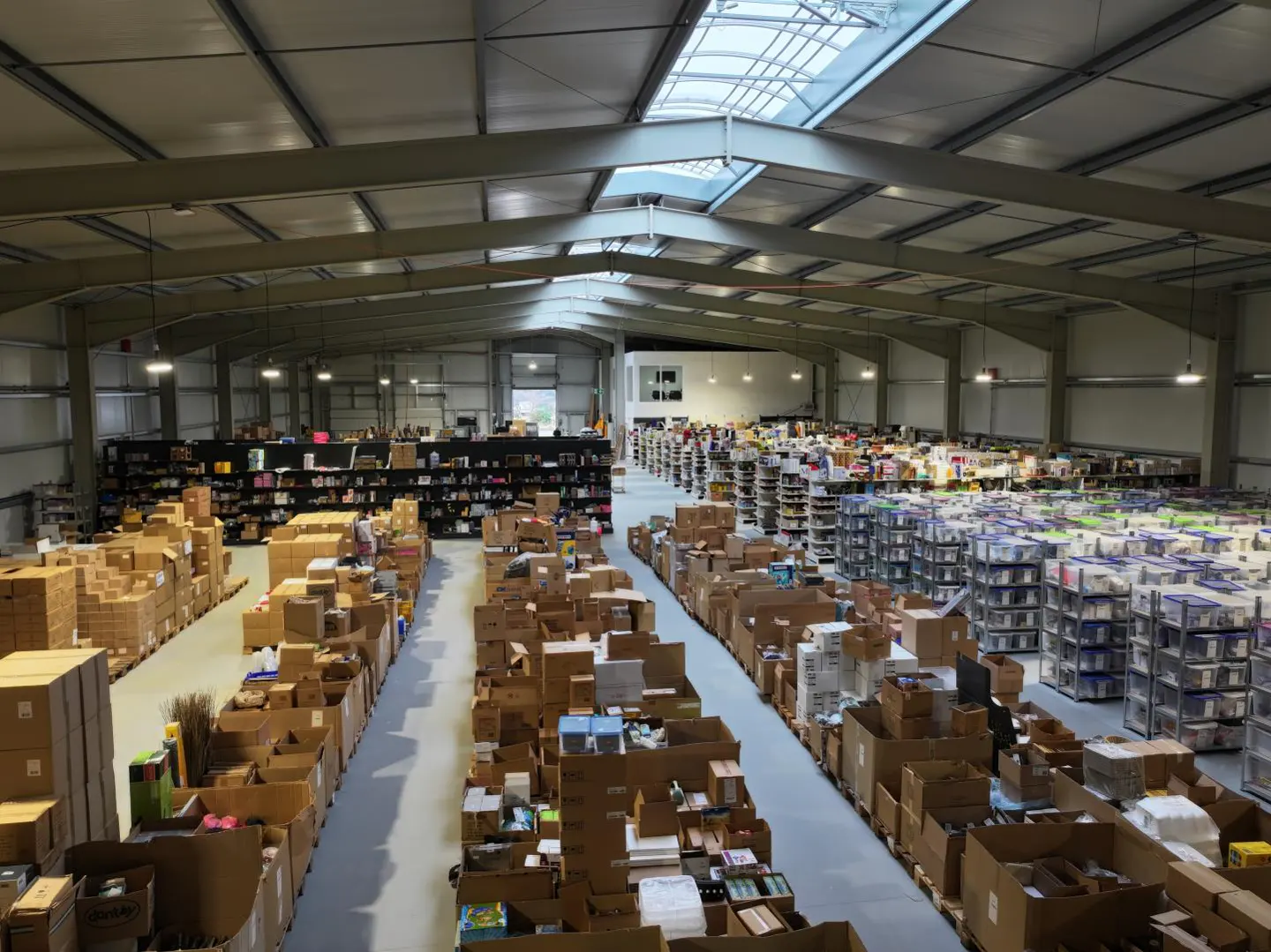 A large warehouse interior with rows of cardboard boxes and shelves filled with items. Skylights provide natural light.