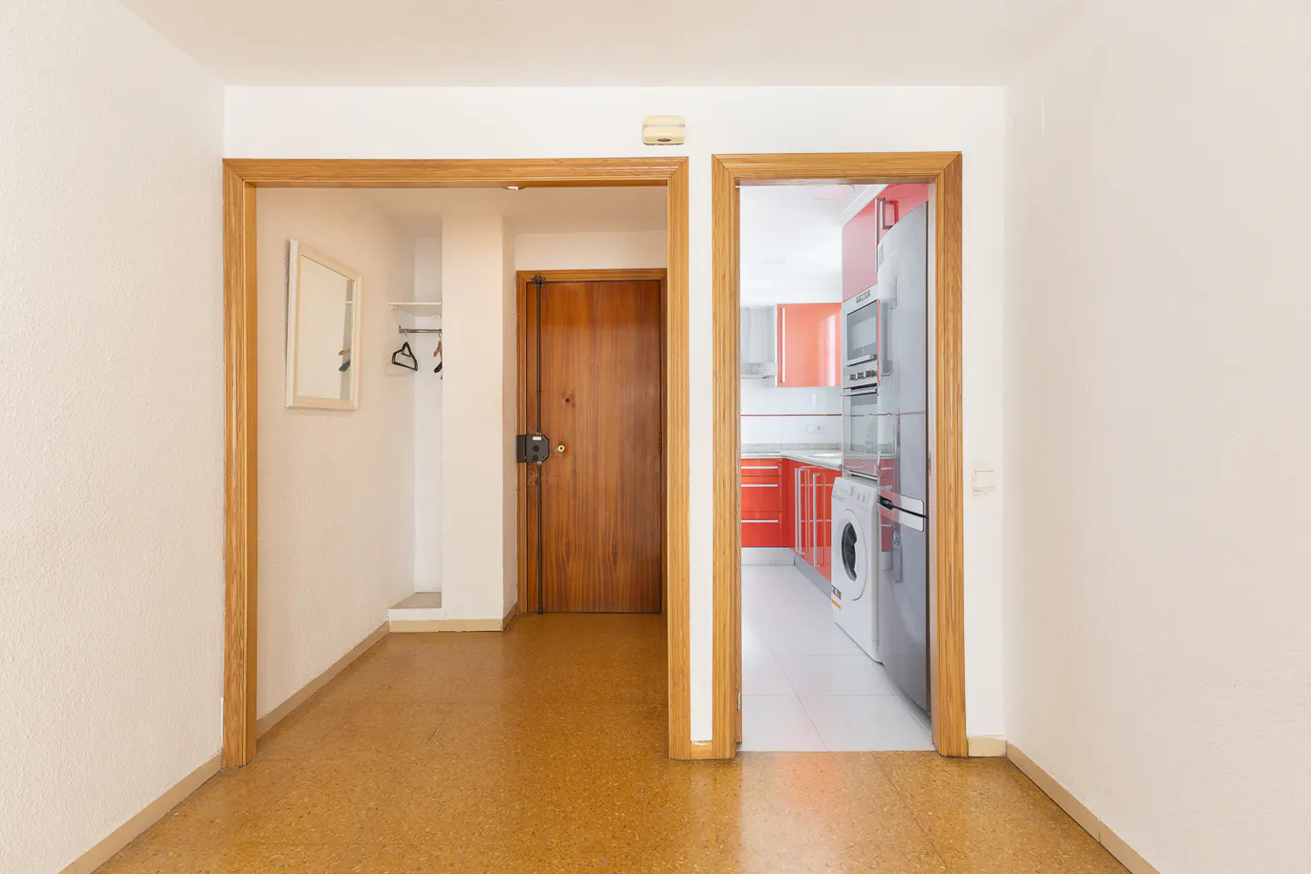 Hallway with wooden door and open doorway leading to a modern kitchen with red cabinets and stainless steel appliances.