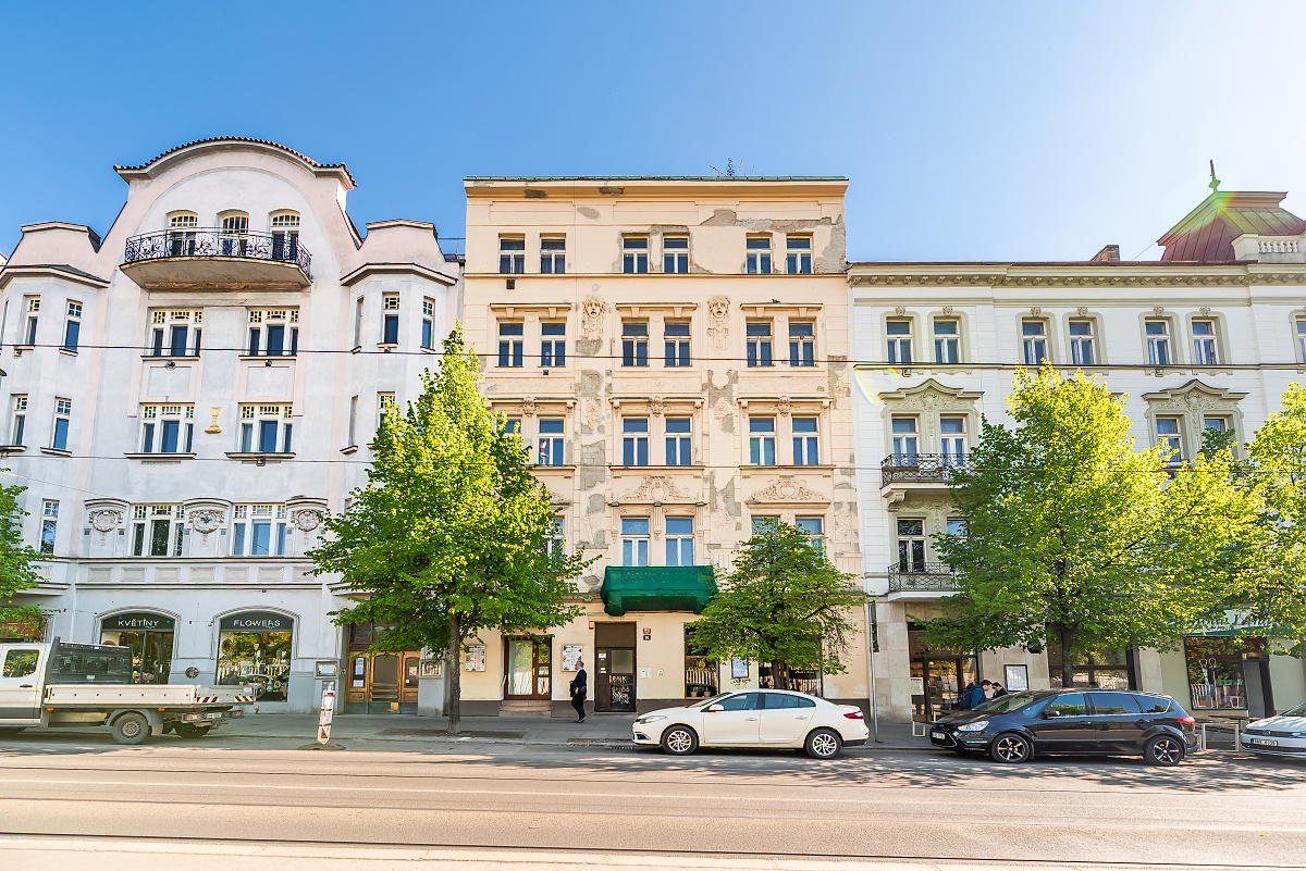 Street view of old European buildings with cars parked along the curb and green trees. Blue sky above.