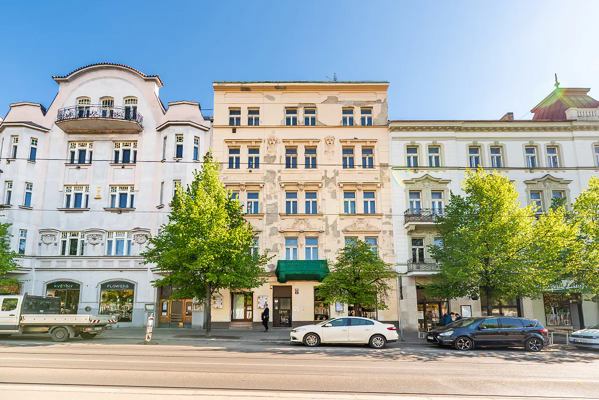 Street view of old European buildings with cars parked along the curb and green trees. Blue sky above.
