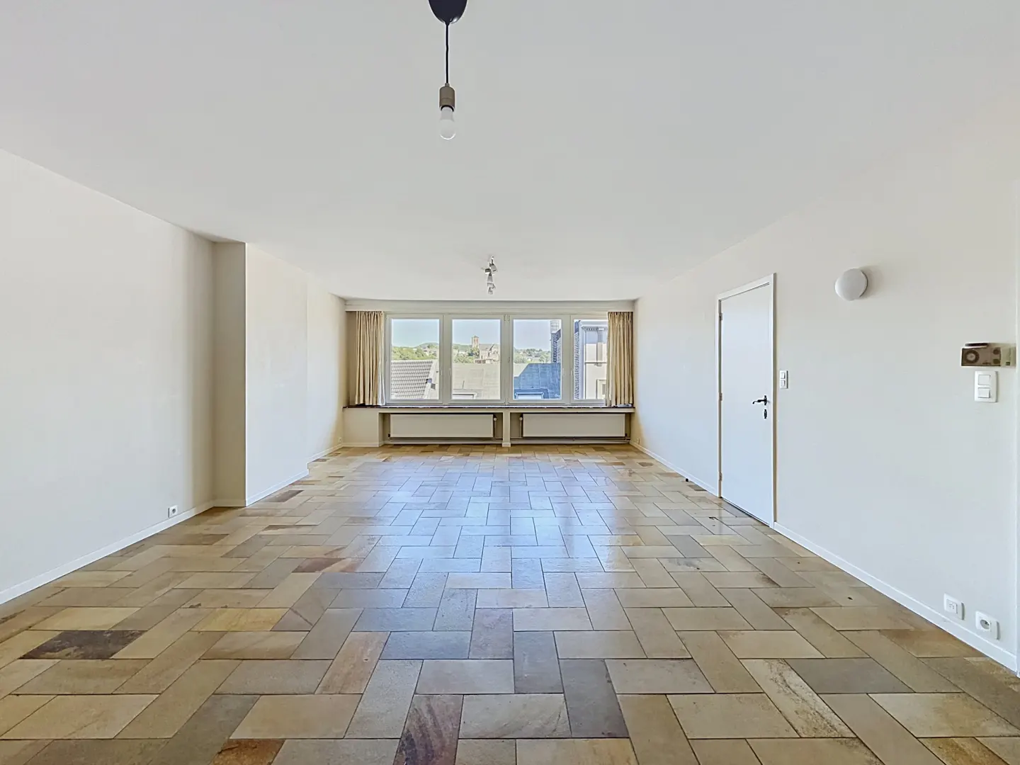 Bright, empty room with herringbone tile floor, white walls, and large windows with tan curtains overlooking a cityscape.