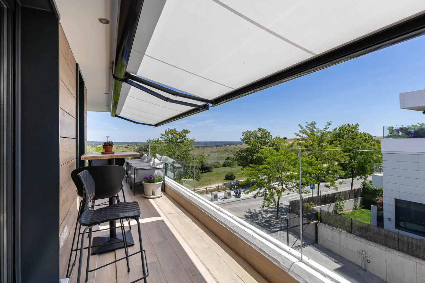 A modern balcony with a retractable awning, black bar stools, and a glass railing overlooking a green landscape.