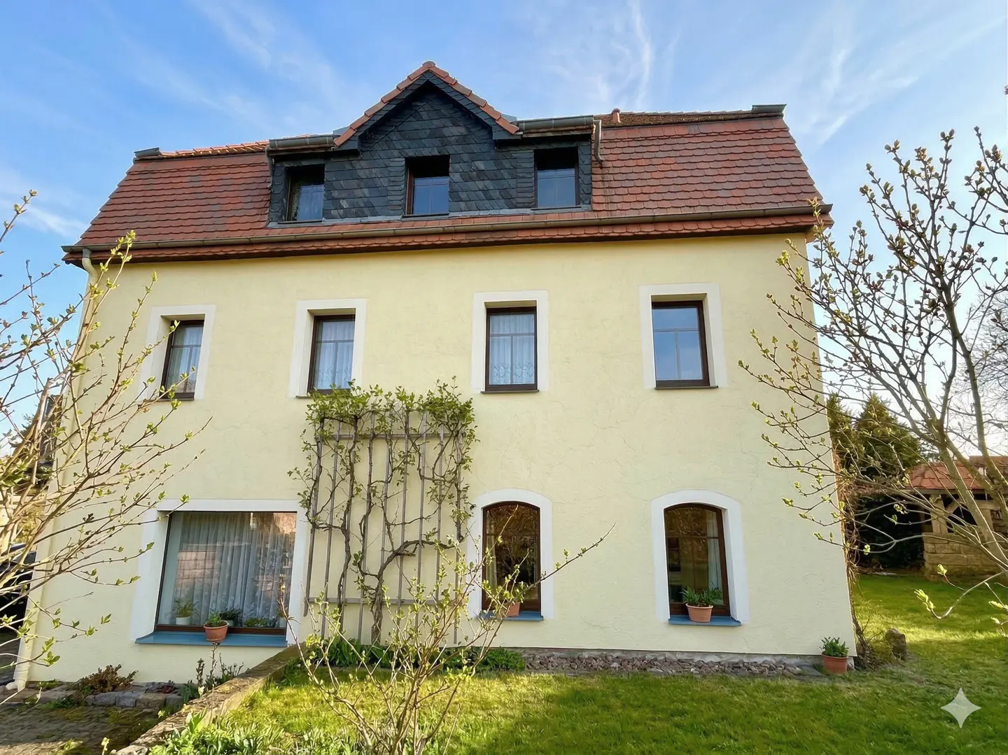 Two-story yellow house with a red tile roof and dark gray dormers. A climbing plant grows on the wall.