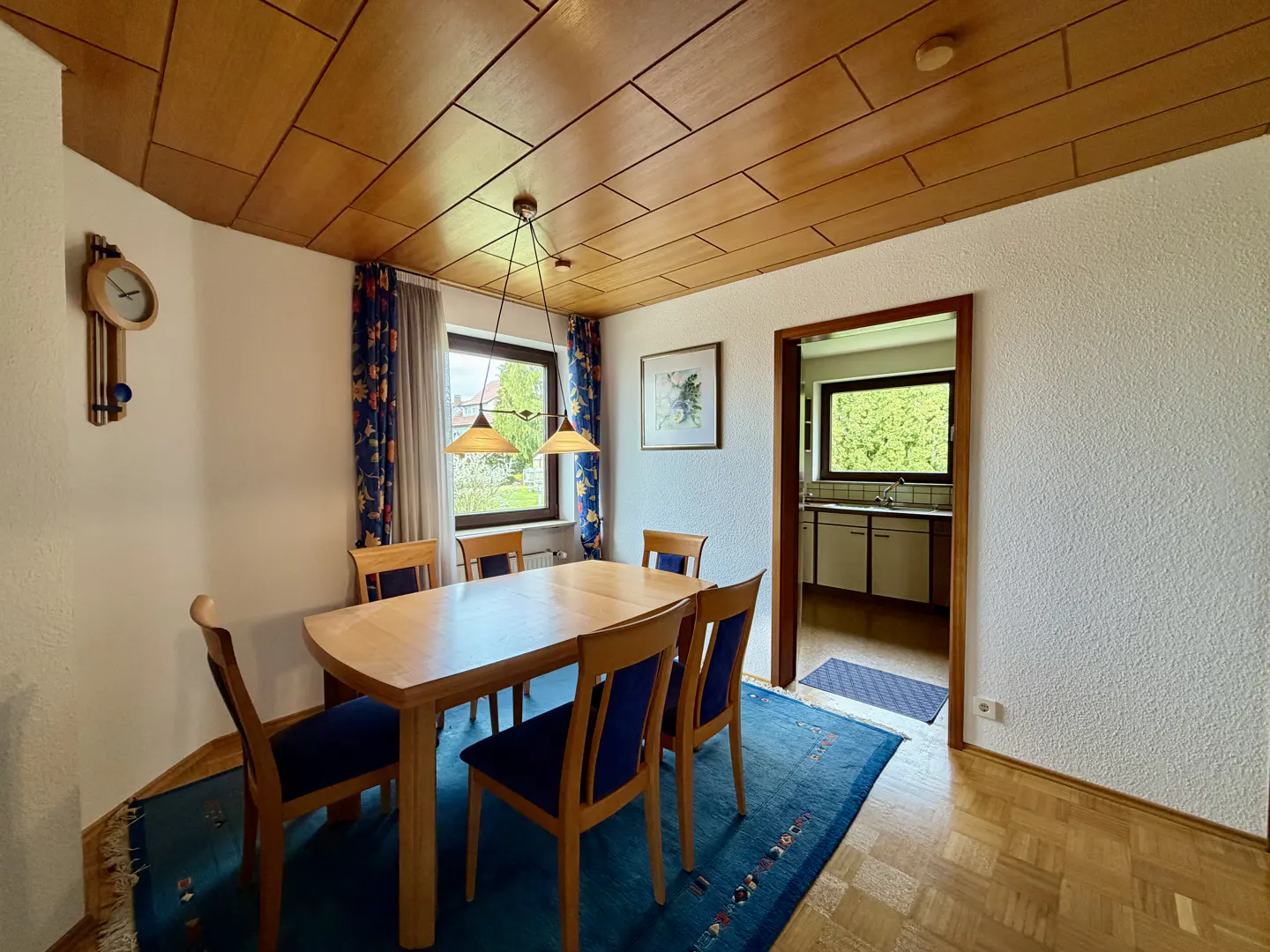 A dining room with a wooden table and blue chairs, a blue rug, and a view into the kitchen.
