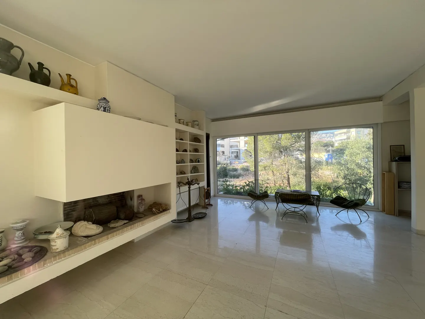 Bright living room with white tile floor, fireplace, and large windows overlooking greenery. Two chairs and a table are near the windows.