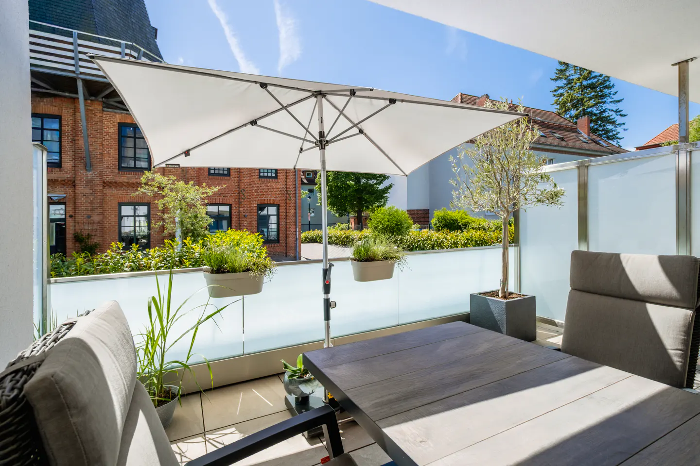 Sunny balcony with a wooden table, grey chairs, a white umbrella, and potted plants overlooking a brick building.